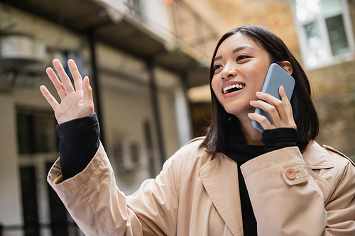 Smiling asian woman talking on smartphone and waving hand outdoors