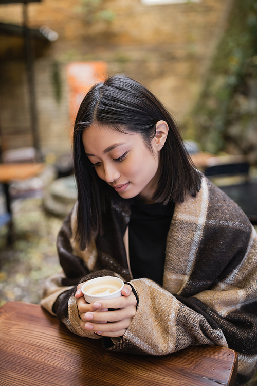 Young asian woman in blanket holding paper cup with cappuccino in outdoor cafe