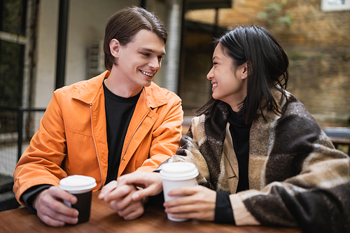 Smiling interracial couple with blanket holding hands near coffee on terrace of cafe