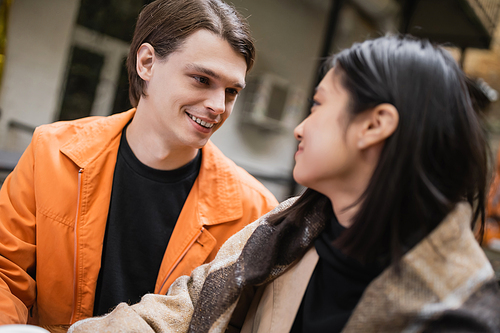 Smiling man looking at blurred asian girlfriend in blanket in outdoor cafe