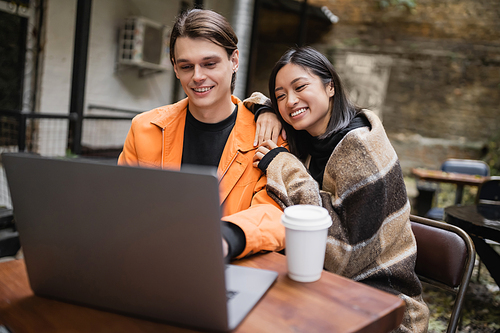 Smiling asian woman in blanket hugging boyfriend near laptop and coffee in outdoor cafe