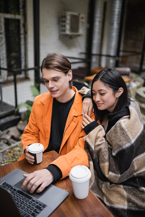 Positive asian woman in blanket hugging boyfriend using laptop near coffee to go on terrace of cafe