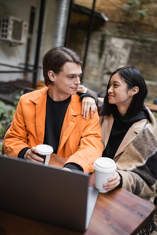 Young interracial couple holding coffee to go and looking at each other near blurred laptop in outdoor cafe