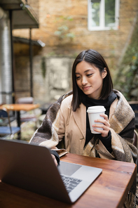 Pretty asian woman in blanket holding coffee to go and using blurred laptop on cafe terrace