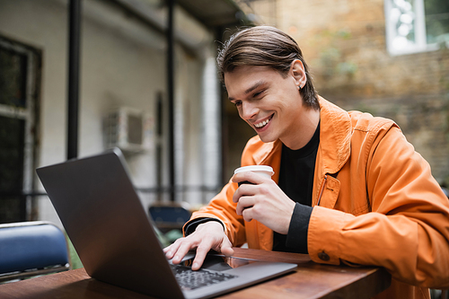 Smiling man holding takeaway drink and using laptop on terrace of cafe