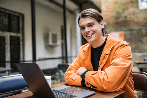 Young freelancer in jacket looking at camera near laptop and coffee to go on terrace of cafe