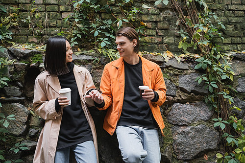 Young multiethnic couple with coffee to go holding hands near wall on urban street