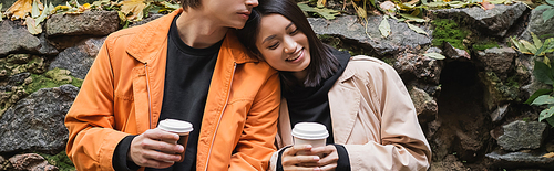 Positive asian woman holding paper cup while standing near boyfriend in jacket on urban street, banner