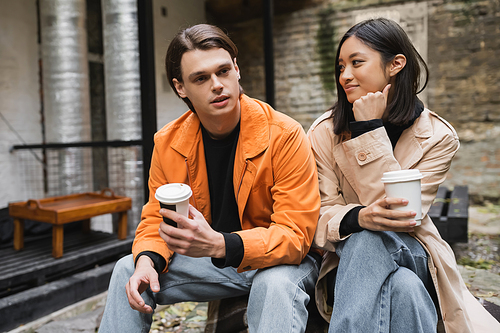 Young man in jacket holding coffee to go and talking to asian girlfriend on terrace of cafe
