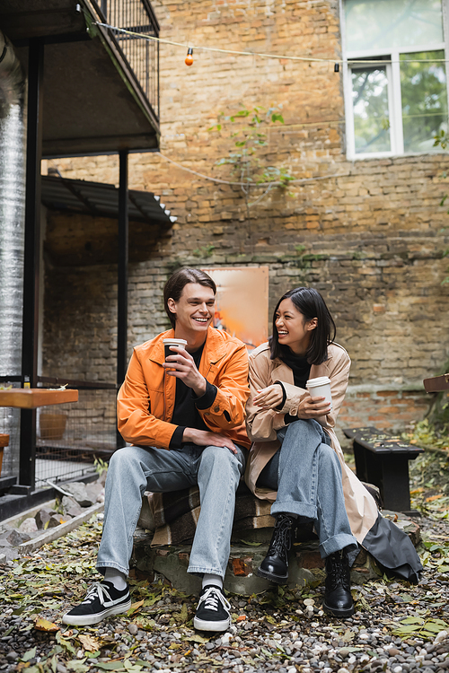 Cheerful and stylish multiethnic couple holding coffee to go on terrace of cafe