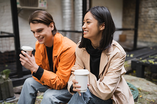 Cheerful asian woman in trench coat holding coffee to go near boyfriend on cafe terrace