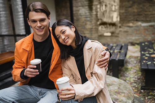 Positive man holding paper cup and hugging asian girlfriend in trench coat in outdoor cafe