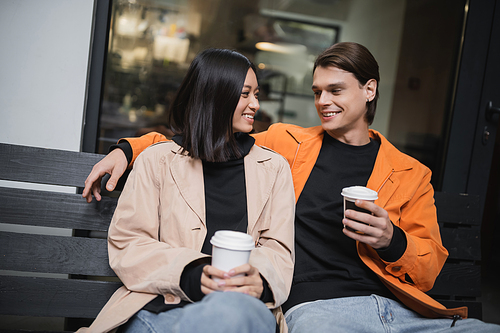 Smiling asian woman holding coffee to go near stylish boyfriend on bench in outdoor cafe