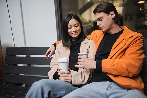 Young interracial couple holding coffee to go while sitting on bench in outdoor cafe
