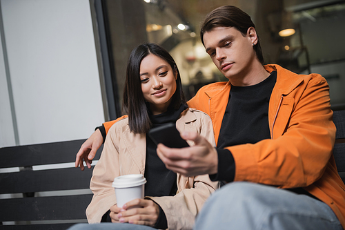 Young multiethnic couple with coffee to go and smartphone sitting on bench on terrace of cafe