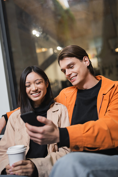 Cheerful multiethnic couple with takeaway drink using smartphone on terrace of cafe