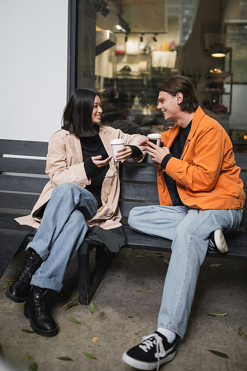 Stylish interracial couple holding coffee to go while talking on bench near cafe