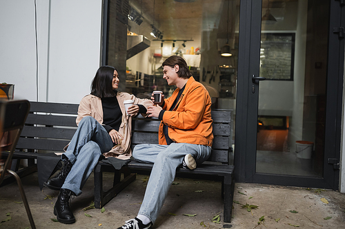 Young multiethnic couple with coffee to go talking on bench on cafe terrace