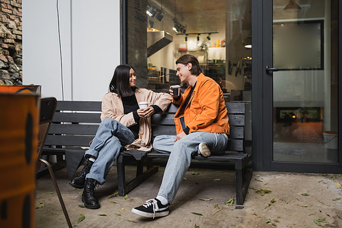 Trendy multiethnic couple talking while holding takeaway cups on bench near cafe