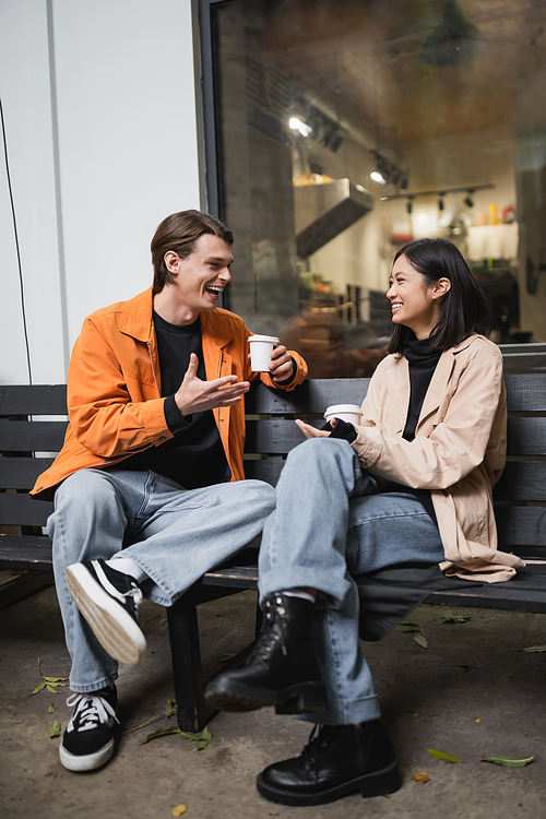 Positive man talking to asian girlfriend with paper cup while sitting on bench near cafe
