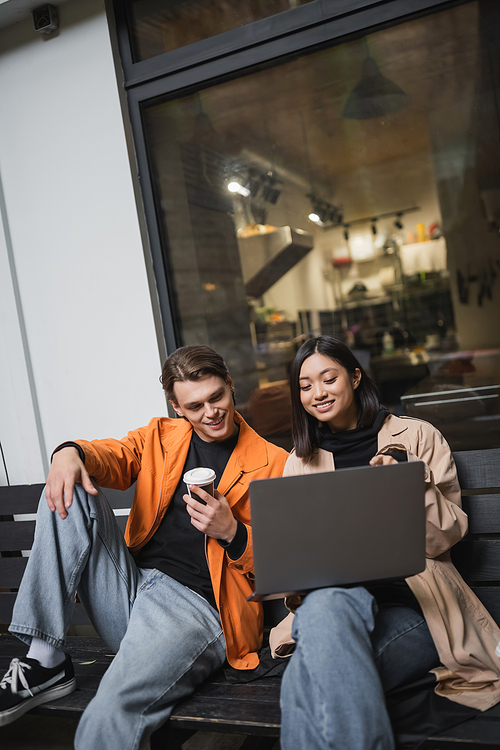 Young multiethnic couple with coffee to go looking at laptop while sitting on bench near cafe