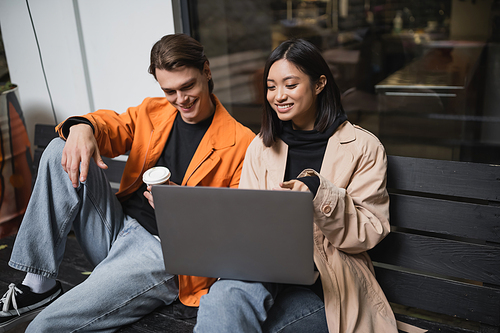 Smiling asian woman pointing at laptop near boyfriend with coffee to go on bench near cafe