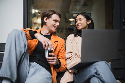 Smiling multiethnic couple with coffee to go and laptop looking at each other near cafe