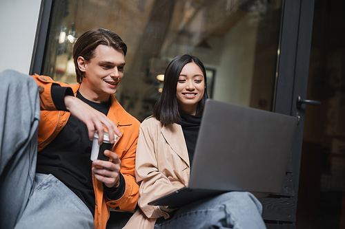 Smiling multiethnic couple with coffee looking at laptop while sitting on bench near cafe