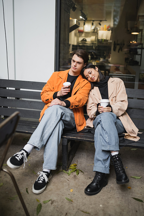 Young asian woman in trench coat holding coffee to go near boyfriend on bench near cafe