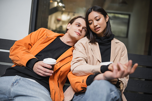 Young interracial couple with coffee to go holding hands on bench near cafe