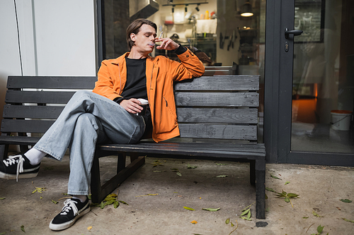 Stylish man smoking cigarette and holding coffee to go near cafe