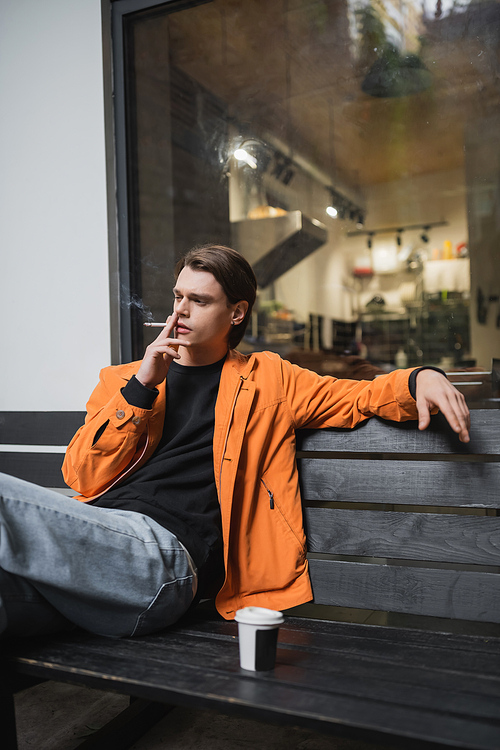 Trendy young man smoking cigarette near coffee to go on bench outdoors