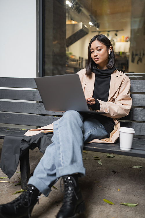 Stylish asian woman in earphone using laptop near paper cup on bench outdoors