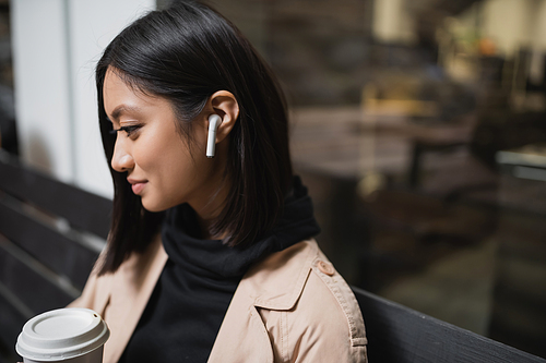 Side view of young asian woman in earphone holding paper cup while sitting on bench near cafe