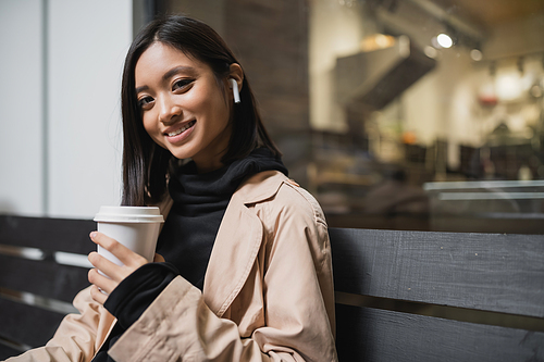 Smiling asian woman in trench coat holding coffee to go and looking at camera on bench near cafe