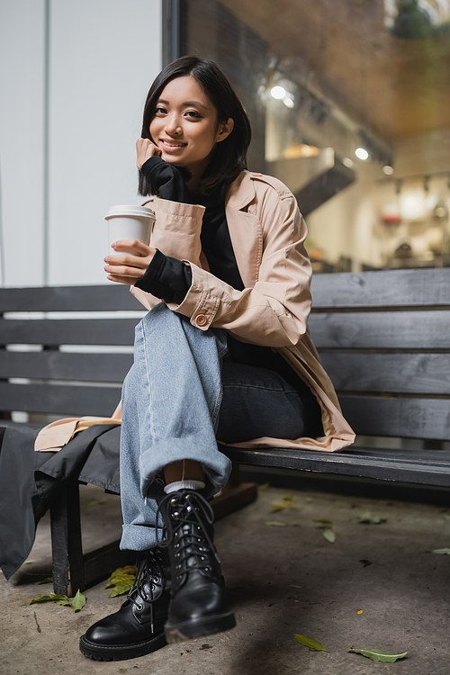 Stylish asian woman in trench coat holding paper cup and looking at camera on bench near cafe