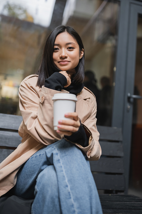 Trendy asian woman in trench coat holding paper cup on bench near cafe