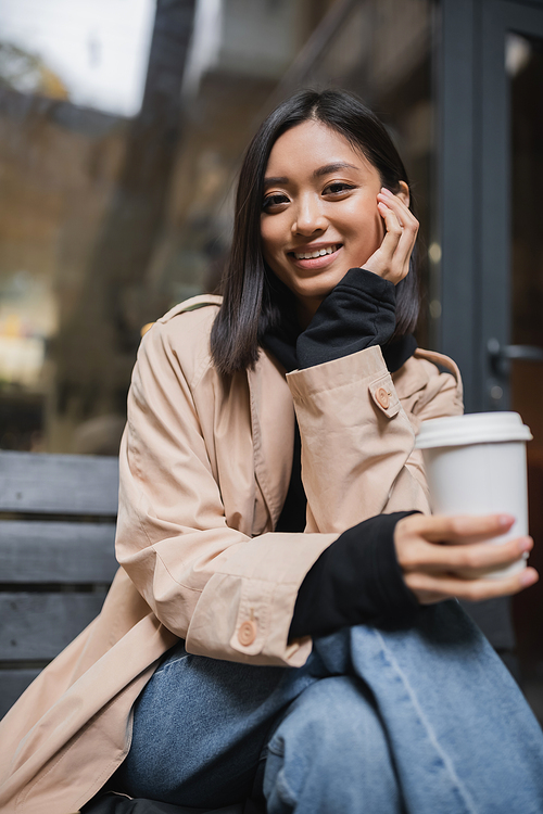 Cheerful asian woman in trench coat holding blurred paper cup and looking at camera near cafe