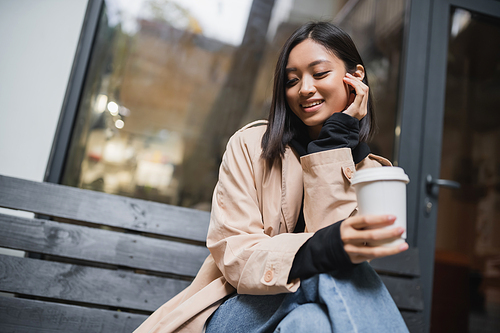Joyful asian woman in trench coat holding takeaway coffee while sitting on bench near cafe