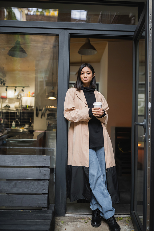 Trendy asian woman in trench coat holding paper cup near cafe