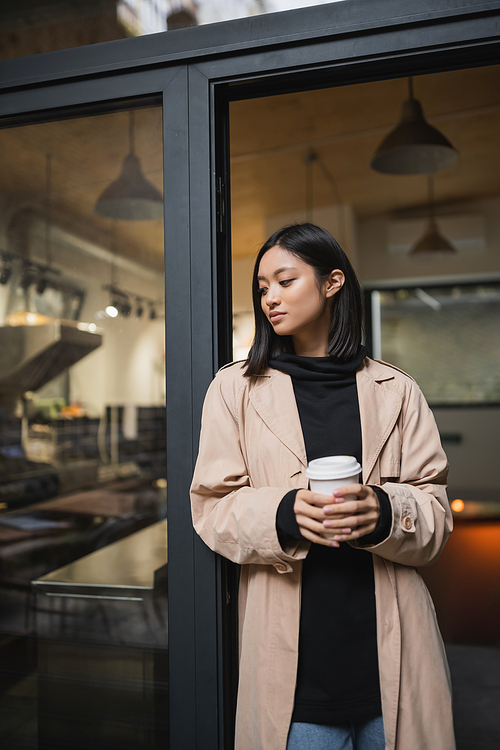 Stylish asian woman in trench coat holding coffee to go near door of cafe