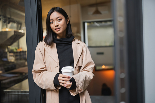 Pretty asian woman in trench coat holding coffee to go near open door of cafe