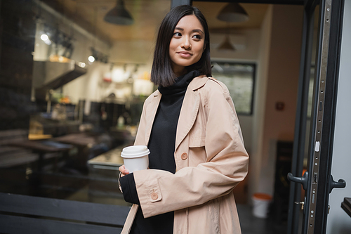 Stylish asian woman holding takeaway coffee near door of cafe outdoors