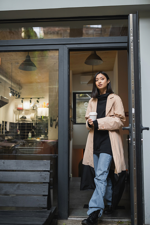 Fashionable asian woman holding coffee to go near open door of cafe