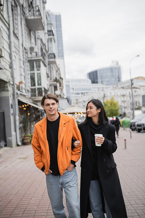 Smiling asian woman holding coffee to go and looking at boyfriend on urban street