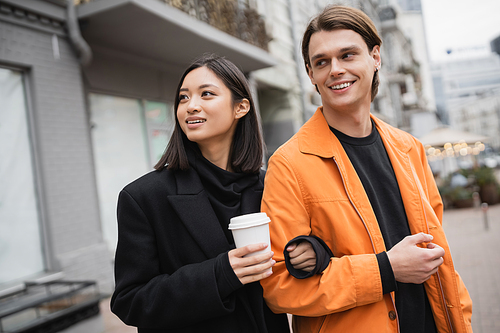 Cheerful asian woman holding paper cup and looking away near boyfriend on street