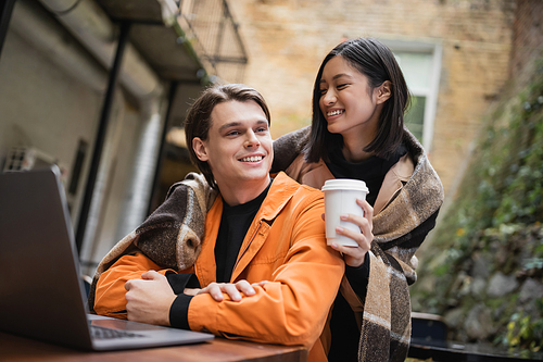 Young asian woman in blanket holding coffee to go and hugging boyfriend near laptop on terrace of cafe
