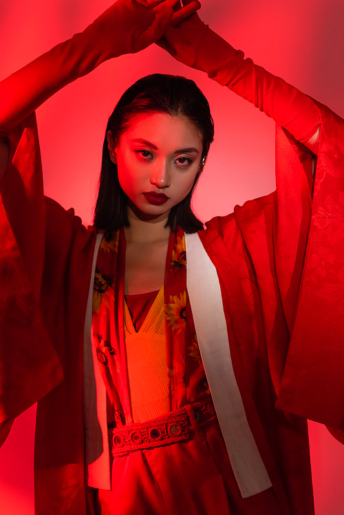 asian woman in kimono cape and gloves posing with hands above head on red background
