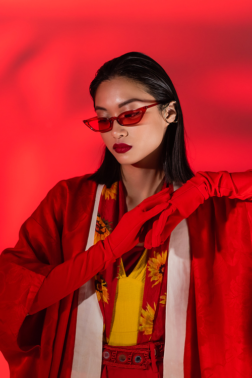 asian model in kimono cape and fashionable sunglasses posing in gloves on background with red shade
