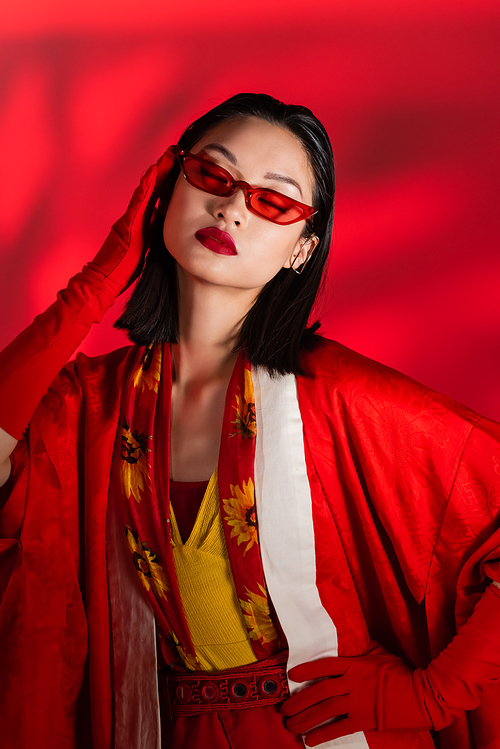 fashionable asian woman in kimono cape and sunglasses posing with closed eyes on red background with shadow